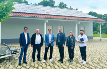 Projektverantwortliche stehen vor einem Gebäude des Parkfriedhofs in Olching, auf dem eine Photovoltaik-Anlage in Betrieb genommen wurde.