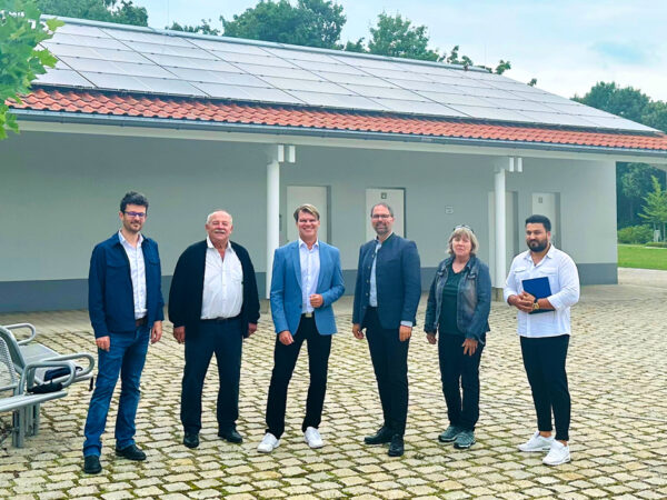 Projektverantwortliche der Stadt Olching und der Stadtwerke Olching stehen vor einem Gebäude des Parkfriedhofs in Olching auf dem eine PV-Anlage installiert wurde.