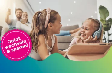 Zwei Mädchen sitzen fröhlich vor einem Tablet. Im Hintergrund sieht man den Vater und die Mutter auf einer Couch im warmer Atmosphäre.