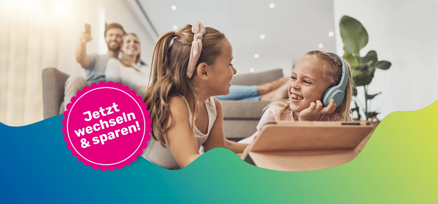 Zwei Mädchen sitzen fröhlich vor einem Tablet. Im Hintergrund sieht man den Vater und die Mutter auf einer Couch im warmer Atmosphäre.