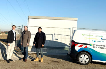20270126_PV Anlage Trafostation Der Geschäftsführer Thomas Grulke, der Erste Bürgermeister von Olching Herr Andreas Magg und der Projektleiter der Stadtwerke Olching Herr Firas Alzaghir stehen auf der Baustelle der geplanten PV-Freiflächenanlage vor einer aufgestellten Trafostation neben einem Fahrzeug der Stadtwerke Olching