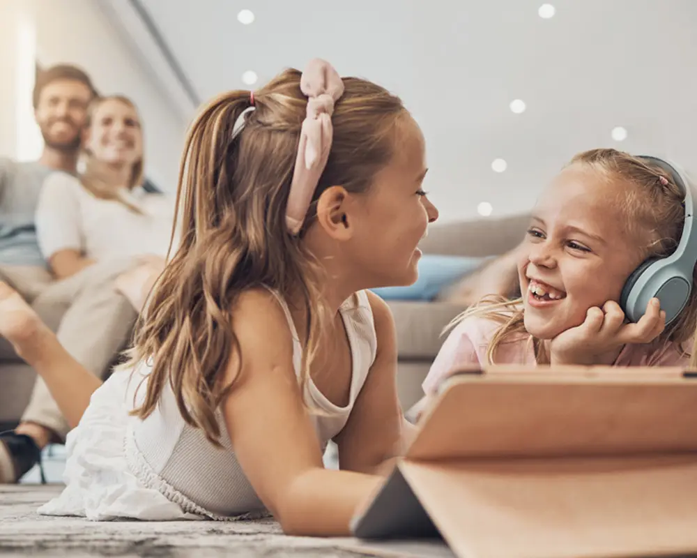 Website_Headerbild_Strom_Startseite Zwei Mädchen sitzen fröhlich vor einem Tablet. Im Hintergrund sieht man den Vater und die Mutter auf einer Couch im warmer Atmosphäre.