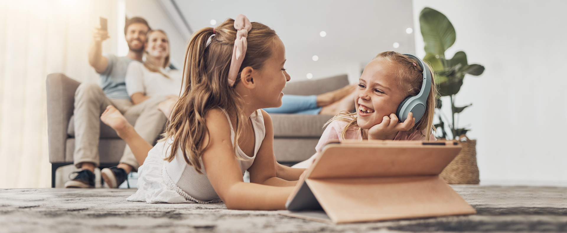 Zwei Mädchen sitzen fröhlich vor einem Tablet. Im Hintergrund sieht man den Vater und die Mutter auf einer Couch im warmer Atmosphäre.